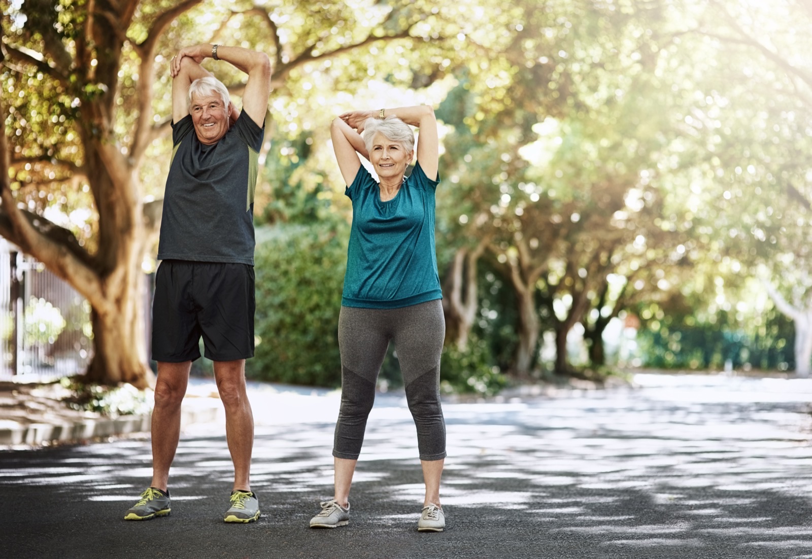 A smiling senior couple stretching together outdoors on a tree-lined path.