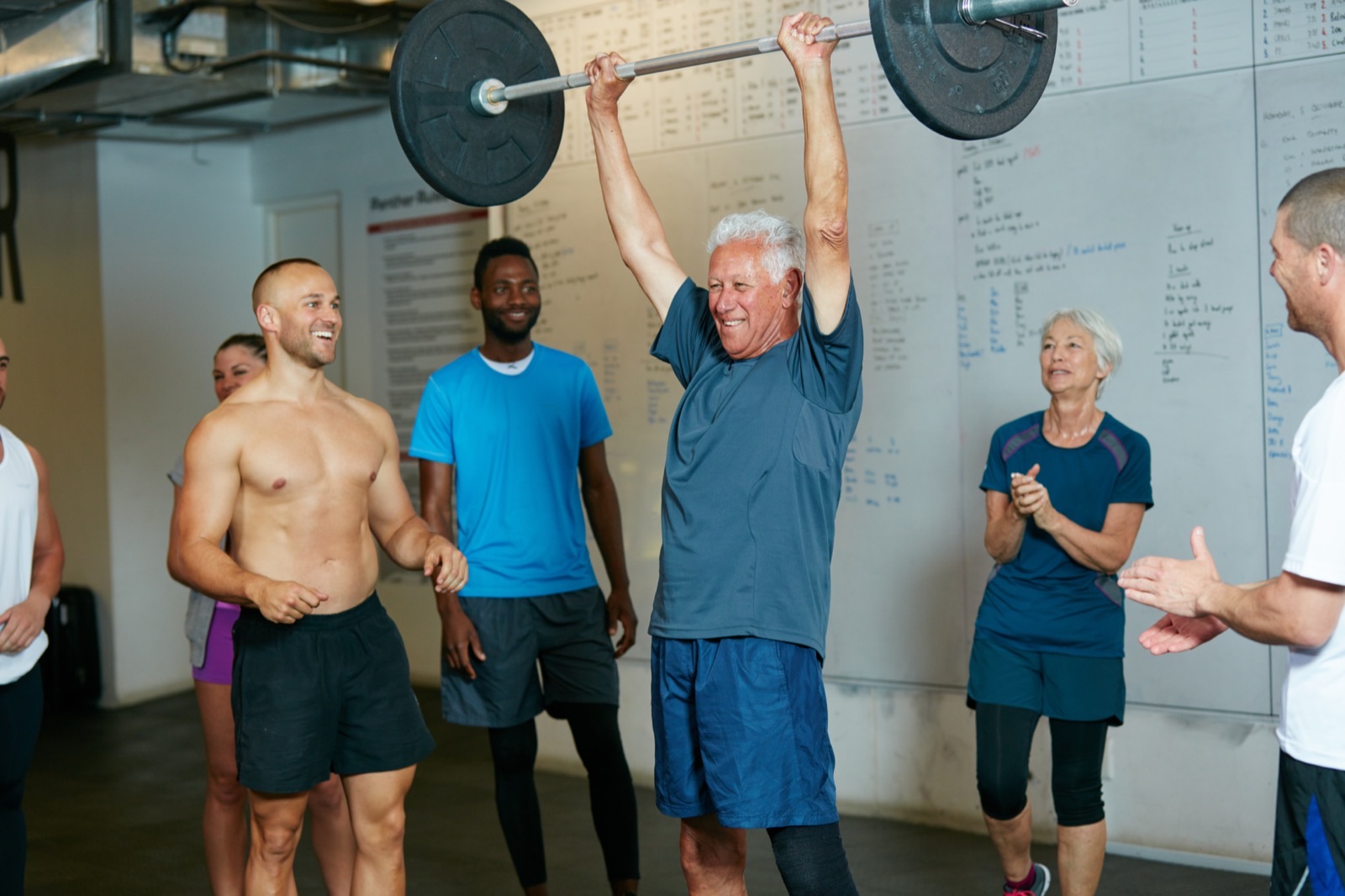 A senior man lifting a barbell overhead at the gym, supported and cheered on by other participants.