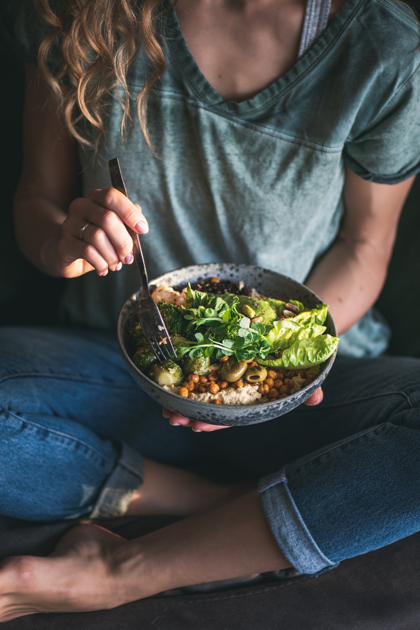 A woman sitting cross-legged, eating a wholefood bowl of vegetables, chickpeas and hummus with a fork.